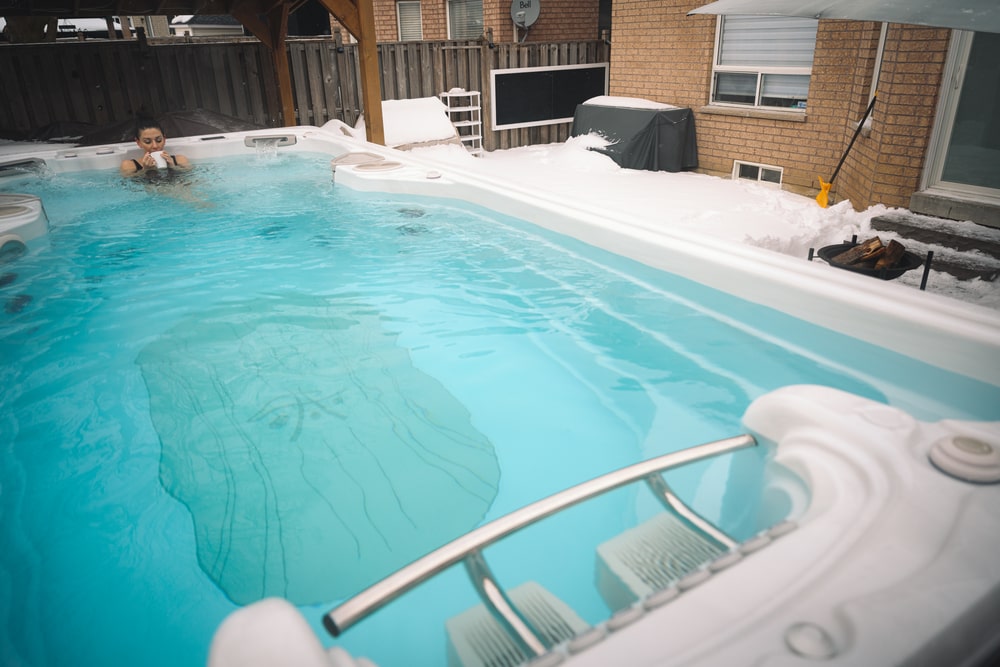 Woman is soaking in a swim spa in a snowy backyard, enjoying her morning coffee